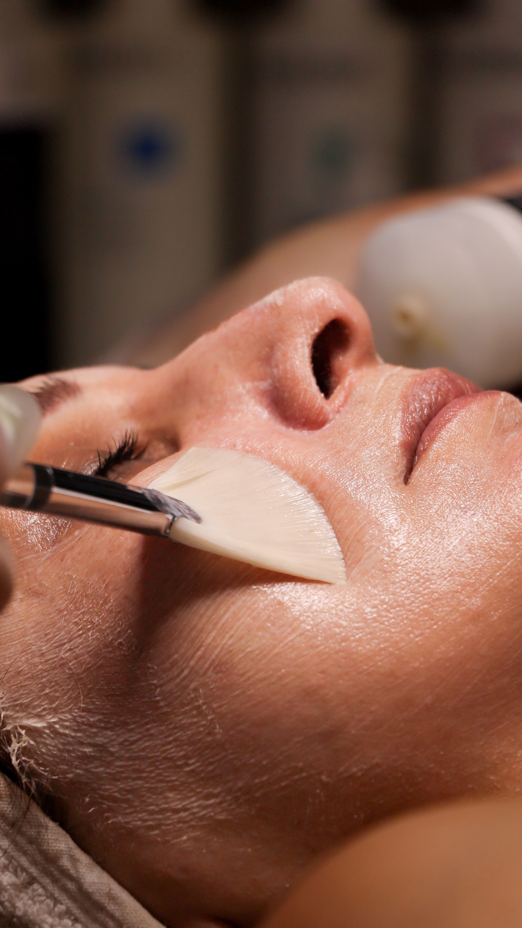 Close-up of a brush sweeping a hydrating mask across a woman's cheek during a facial